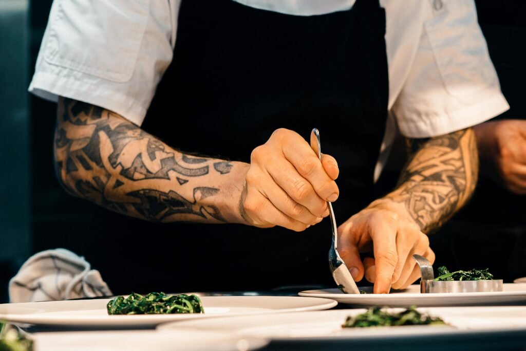 A tattooed chef carefully plates a spinach dish in a professional kitchen setting, showcasing culinary expertise.